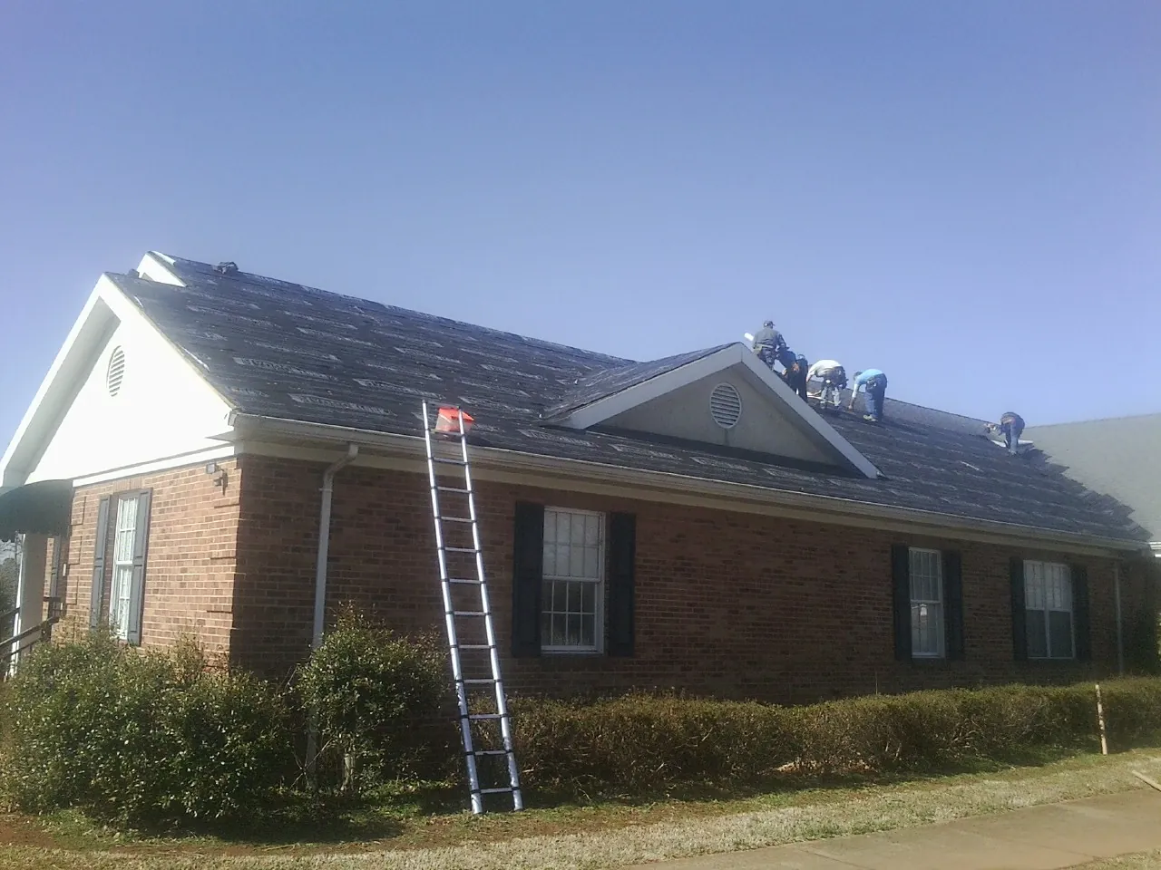 Crew tearing shingles off a brick ranch roof with a ladder set — Emergency Roof Repair work by our crew in Eudora